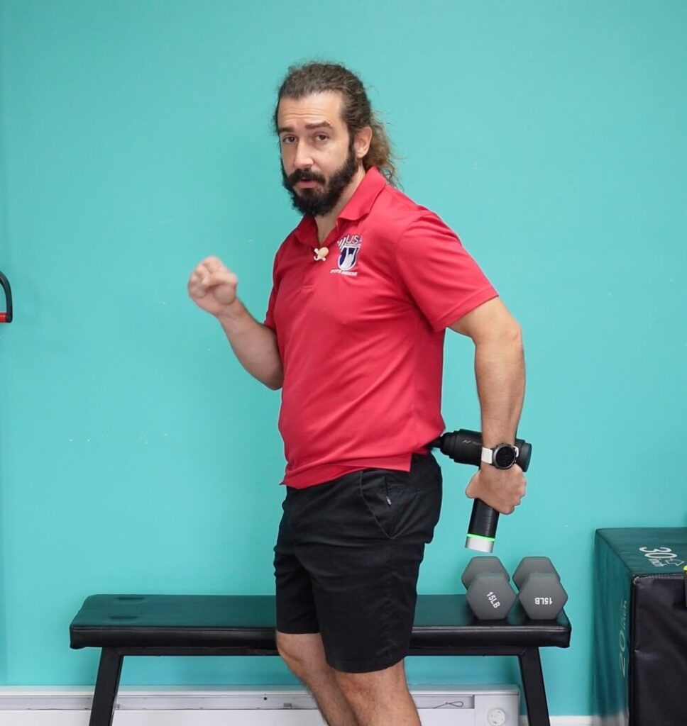 Dr. john standing holding the percussion therapy gun in his left hand using it to massage his low back for back pain relief